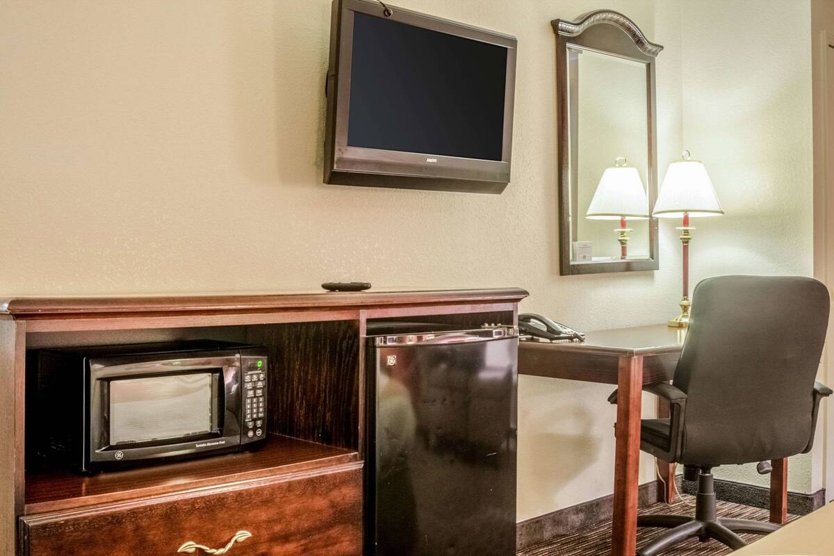 A hotel room with a microwave, a refrigerator, a desk, a chair, a television, and a mirror.