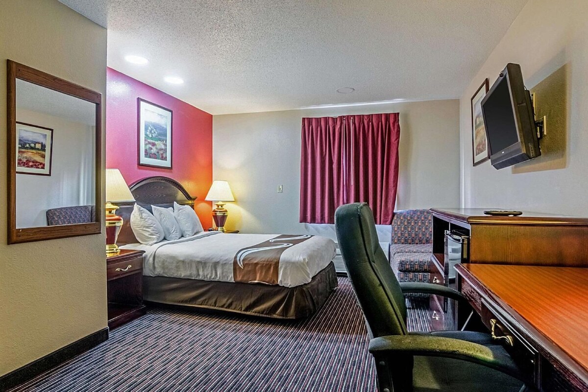 A hotel room with a bed, a desk, a chair, a TV, and a window with curtains.