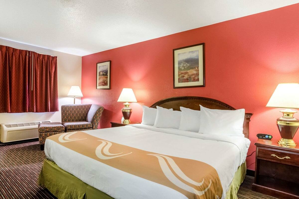 A hotel room with a large bed, two bedside lamps, a chair, a heater, and two framed pictures on the wall.