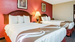 A hotel room with two beds, each with white linens and brown decorative pillows. There is a nightstand with a lamp between the beds. The walls are painted red, and there are framed pictures on the wall.