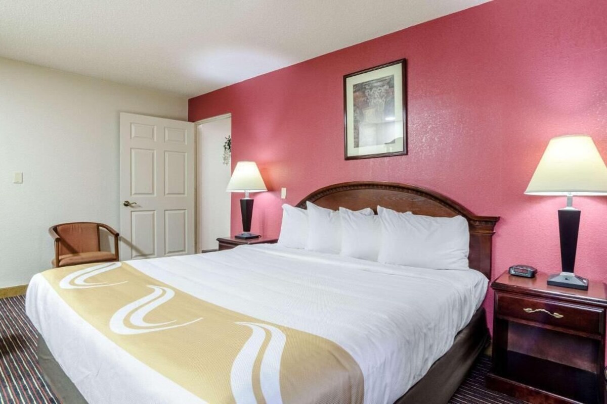 Une chambre d’hôtel avec un lit, deux lampes de chevet, une chaise, une porte et un tableau encadré au mur.