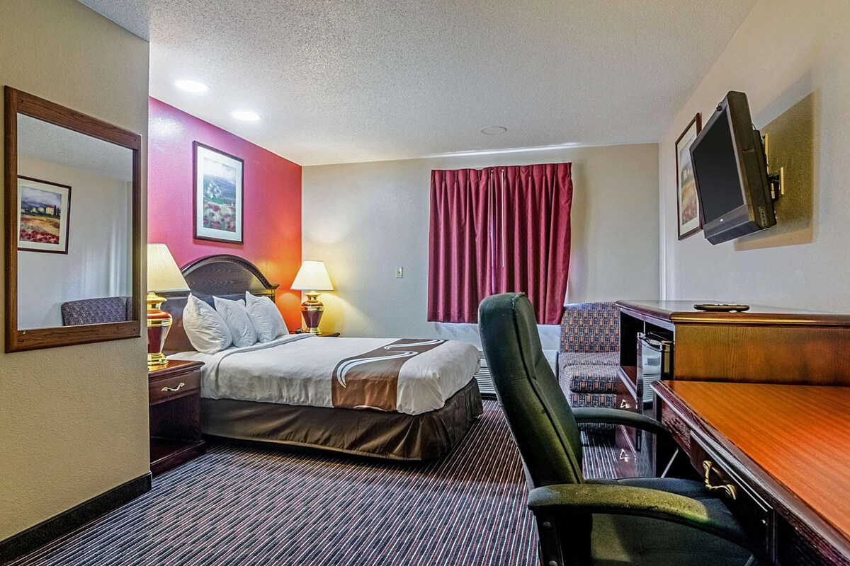 A hotel room with a bed, a desk, a chair, a TV mounted on the wall, and a window with curtains.