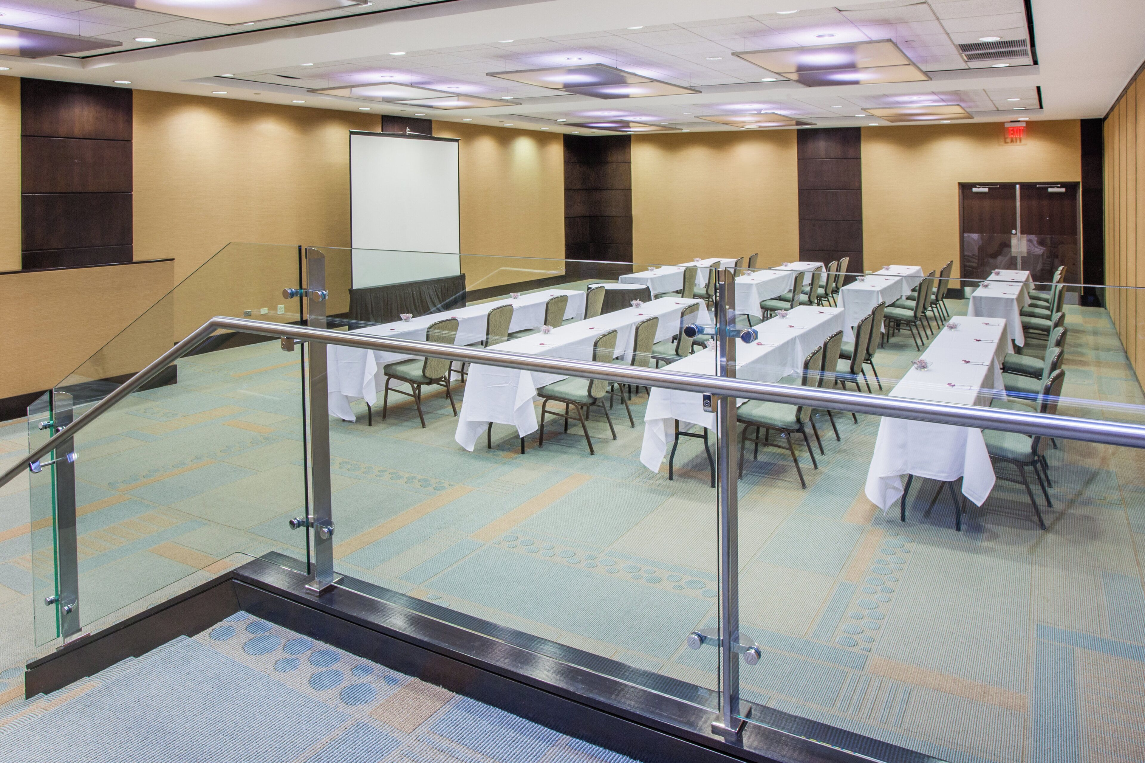 A conference room with multiple tables and chairs, a projector screen, and a staircase with glass railings.