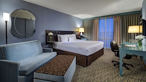 A hotel room with a bed, a sofa, a desk, a chair, a lamp, a mirror, and a window with curtains.