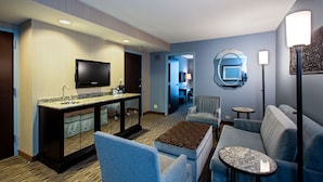 A hotel room with a kitchenette, a flat-screen TV, a sofa, a coffee table, and a round mirror.
