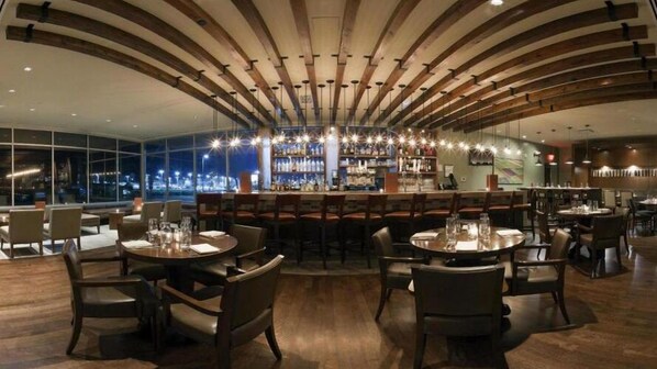 A modern restaurant with a bar area, round tables, and chairs, featuring a unique ceiling design and large windows. - Crowne Plaza Lombard Downers Grove by IHG (Glen Ellyn)