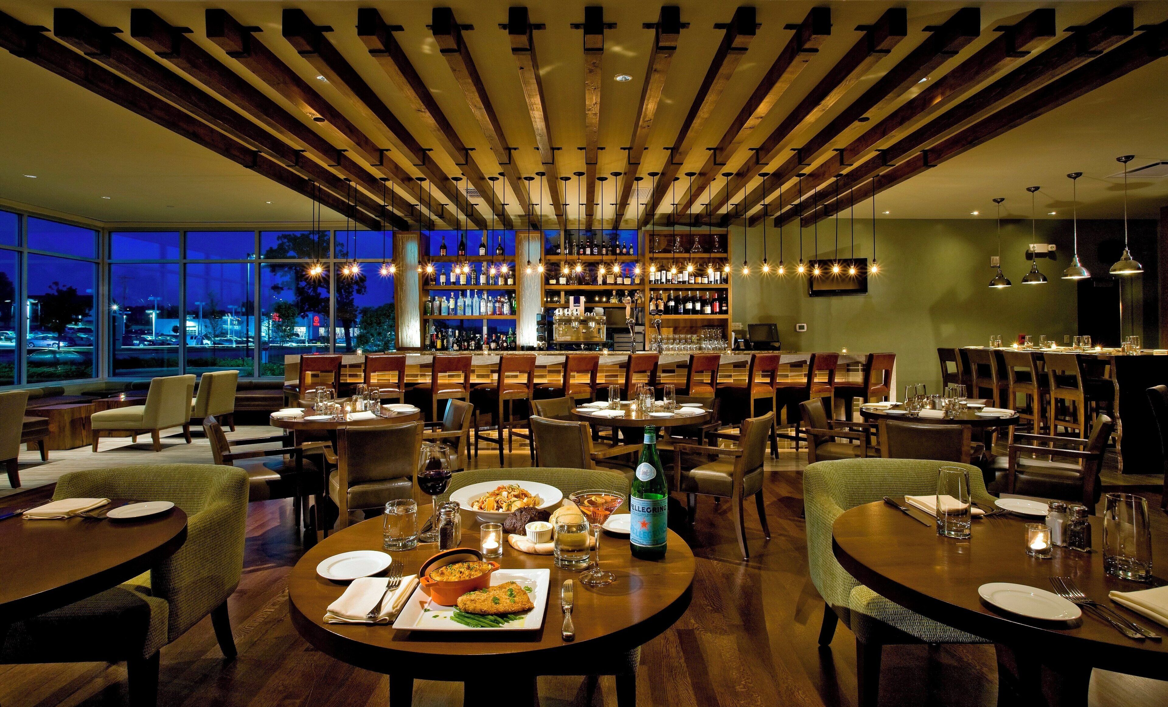 Un restaurant avec des tables rondes, des chaises, un bar avec des étagères remplies de bouteilles, et une vue sur le paysage urbain grâce à de grandes fenêtres.
