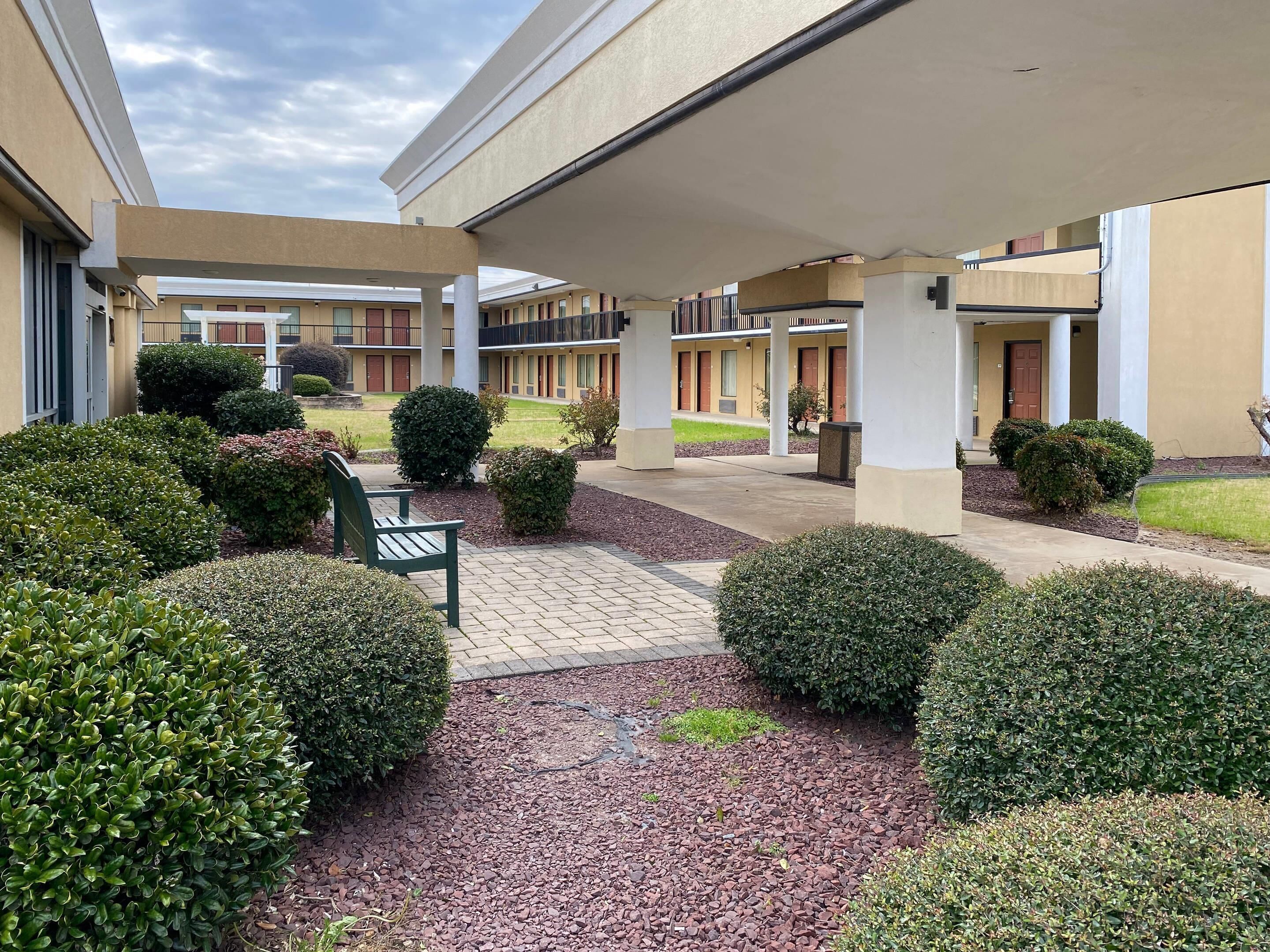 A courtyard with a covered seating area, neatly trimmed bushes, and a pathway leading to a building with multiple doors.