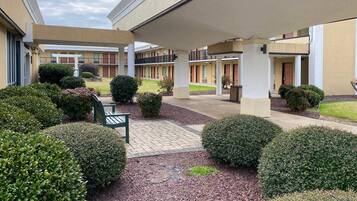 A courtyard with a covered seating area, neatly trimmed bushes, and a pathway leading to a building with multiple doors.