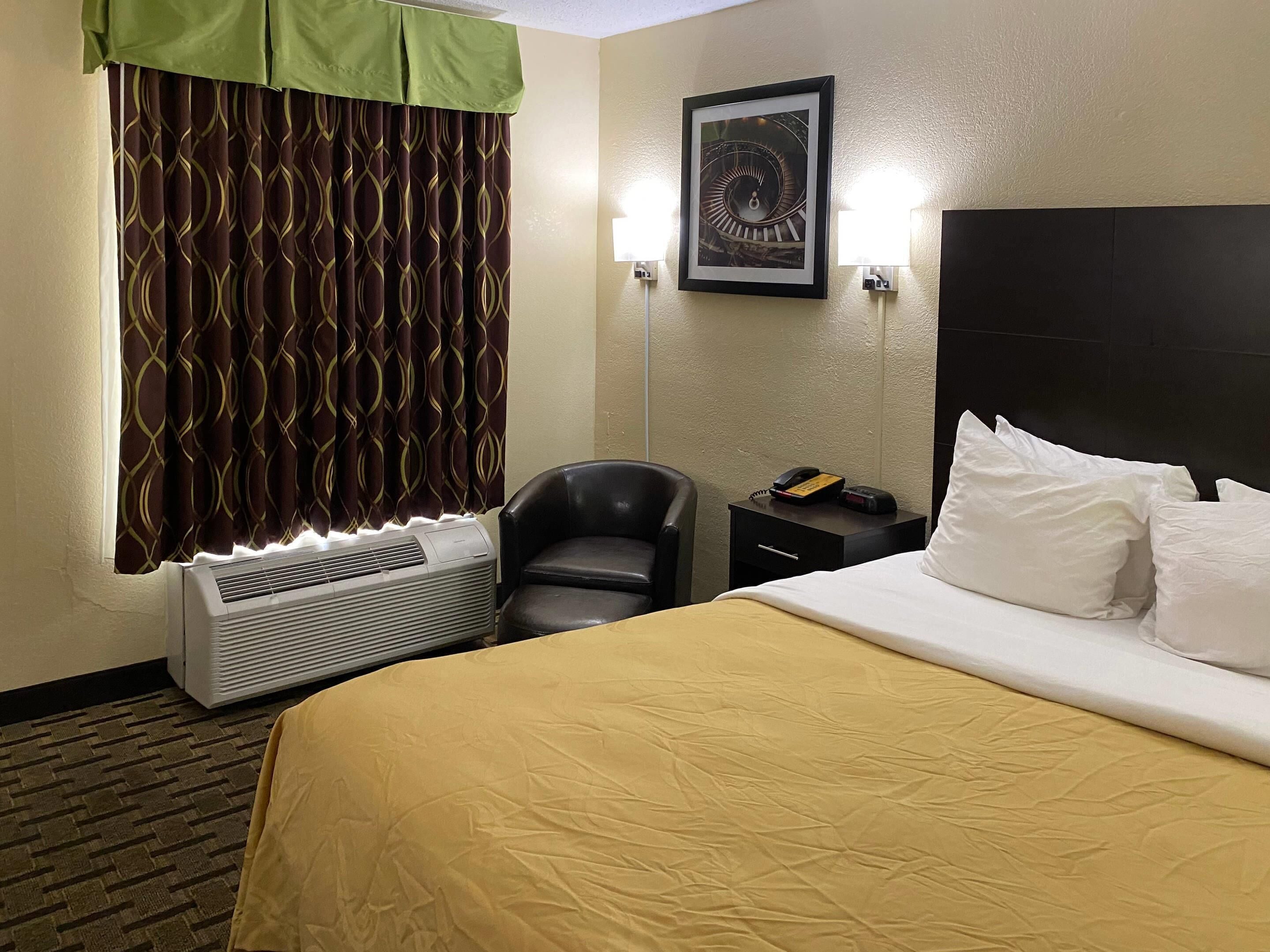 A hotel room with a bed, a chair, a bedside table with a phone, a wall-mounted picture, and a window with patterned curtains.