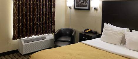 A hotel room with a bed, a chair, a bedside table with a phone, a wall-mounted picture, and a window with patterned curtains.