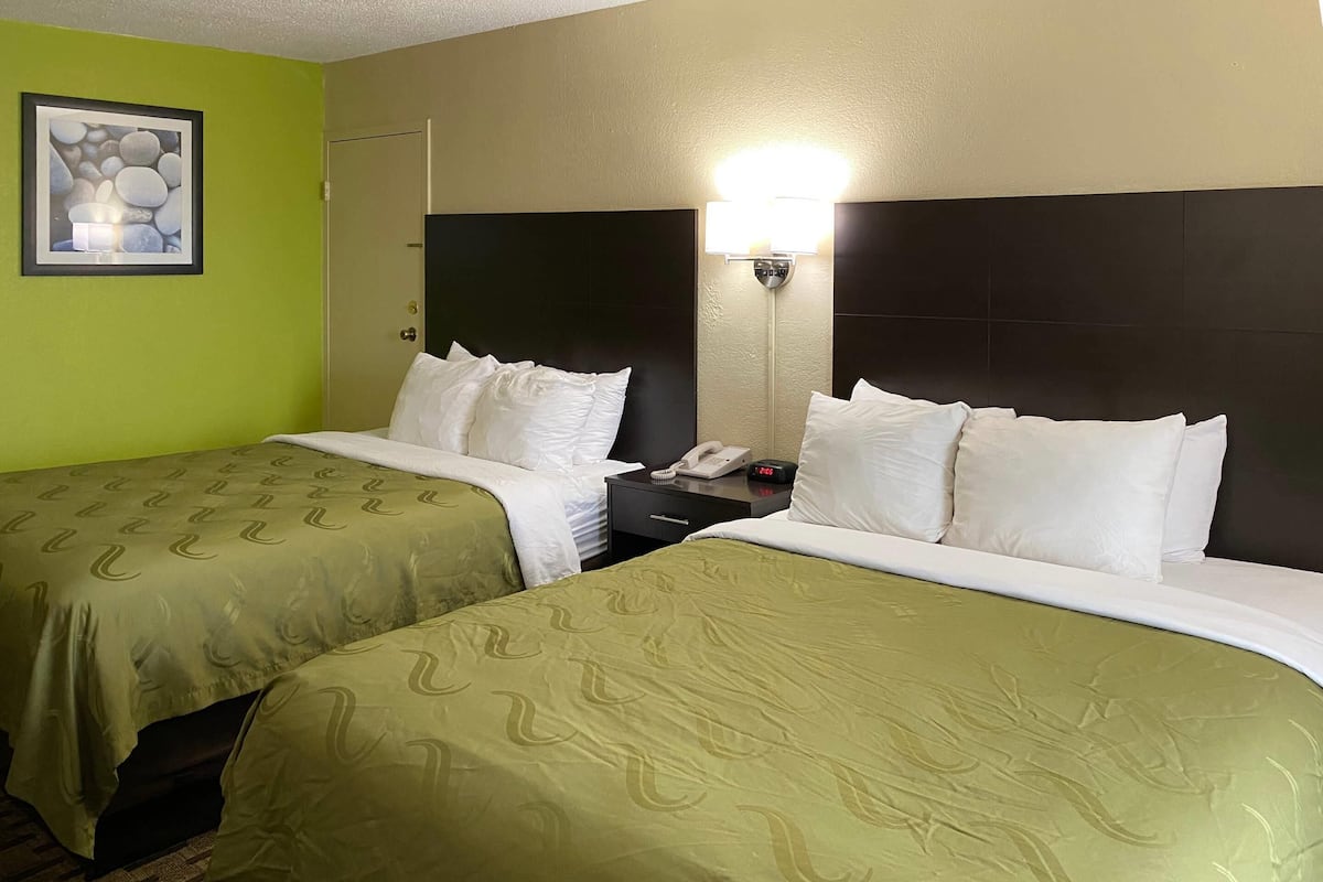 A hotel room with two beds, green bedding, a nightstand with a phone and alarm clock, a framed picture on the wall, and a light fixture above the nightstand.