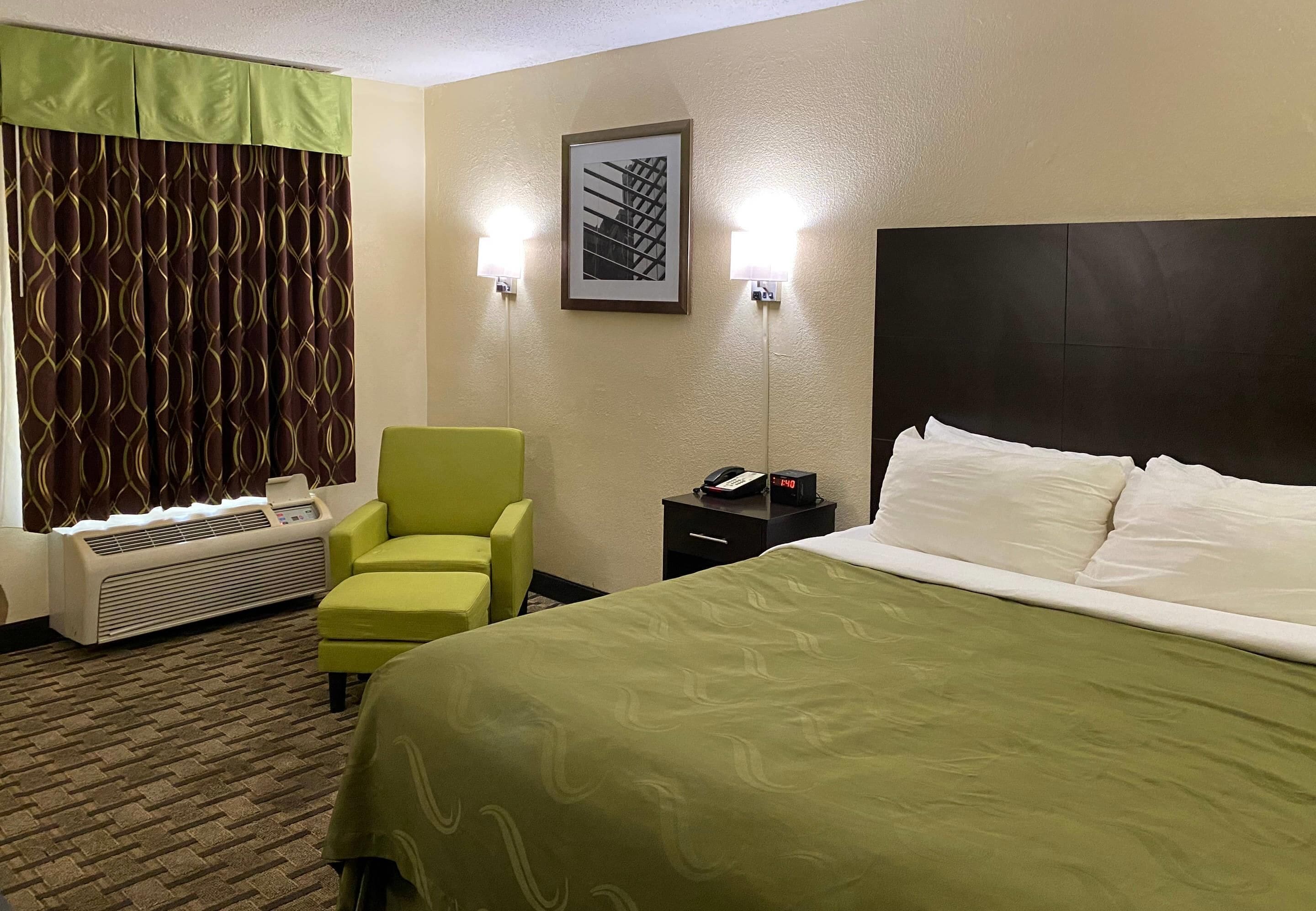 Une chambre d’hôtel avec un lit vert, un fauteuil vert, un radiateur, une table de chevet avec un téléphone et un tableau encadré au mur.