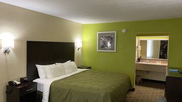A hotel room with a bed, bedside table with a phone, a nightstand, a green wall, a framed picture, a mirror, and a bathroom with a sink and toiletries.