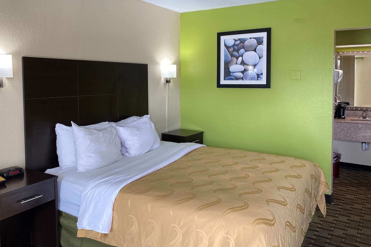 A hotel room with a bed, a nightstand, a framed picture on the wall, and a bathroom visible through an open door.