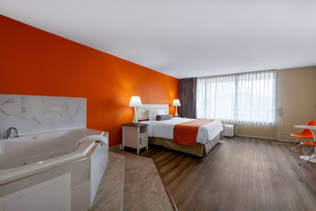 A hotel room with a large bed, a separate bathroom with a bathtub, a desk with a chair, and a window with curtains.