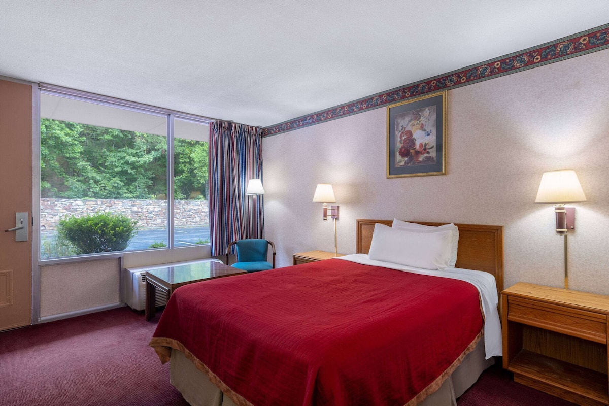 A hotel room with a large bed, two bedside lamps, a chair, a window with curtains, and a painting on the wall.