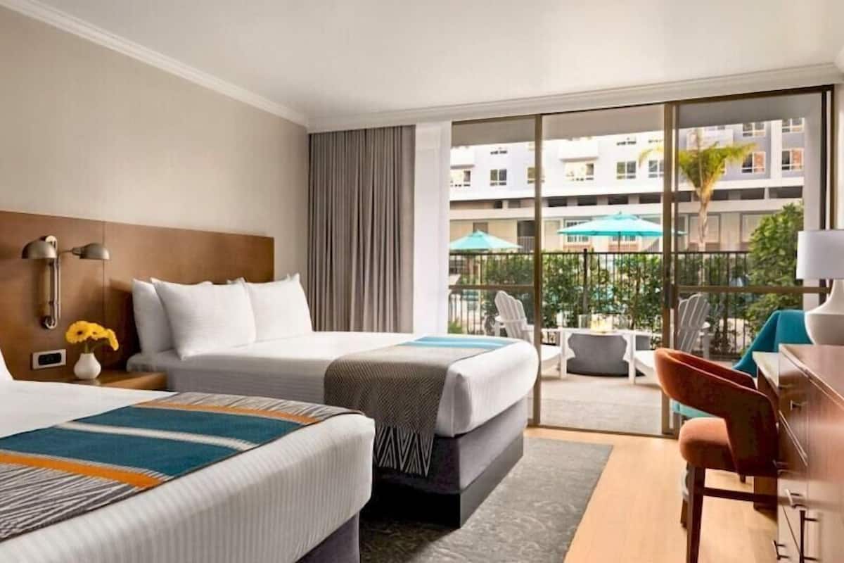 A hotel room with two beds, a desk, a chair, a TV, and a balcony with a view of buildings and greenery.