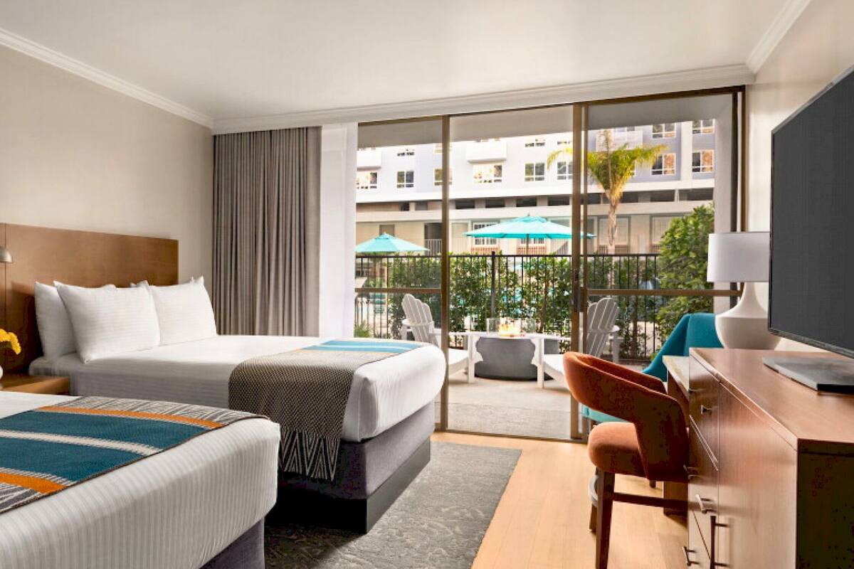 A hotel room with two beds, a desk, a chair, and a balcony with a view of the city.