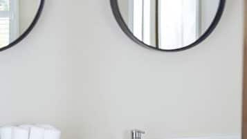 A bathroom with two round mirrors, a white sink, a silver faucet, a wicker basket with white towels, and a small white box with toiletries.