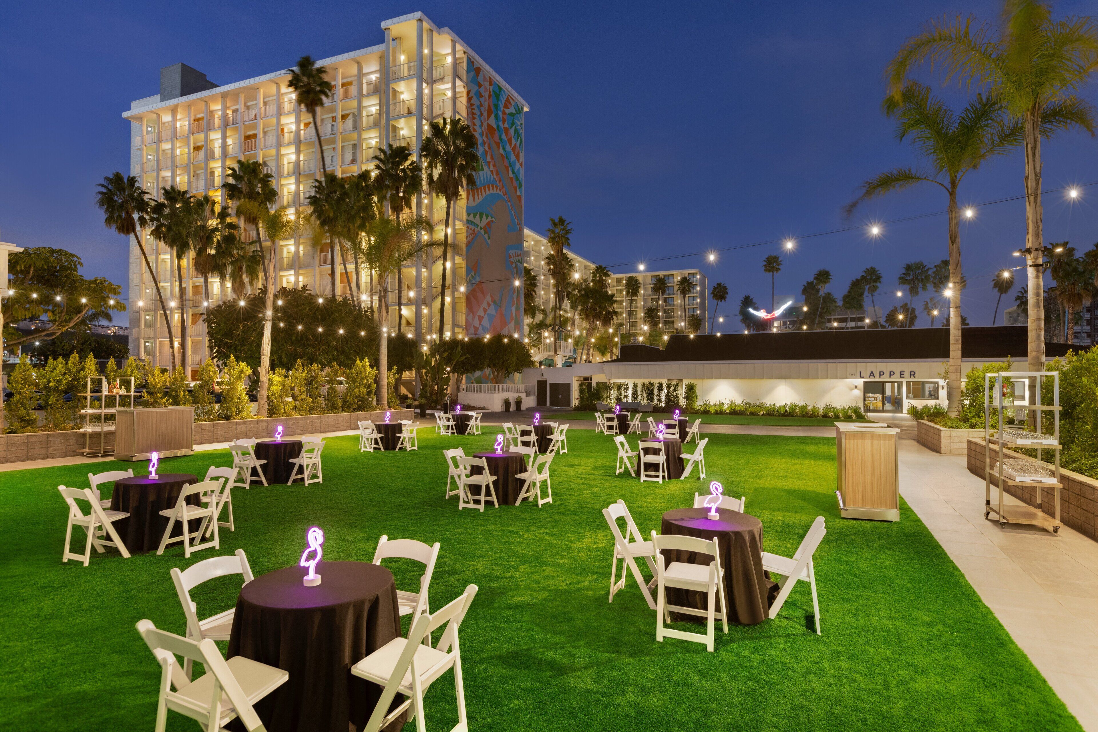 An outdoor dining area with round tables and white chairs, surrounded by palm trees and a building with a mural.