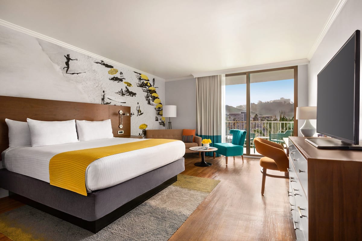 A hotel room with a large bed, a TV, a desk, and a balcony with a view.