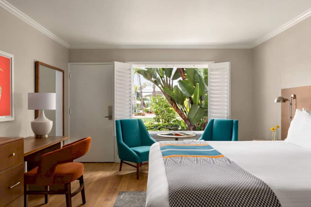 A hotel room with a bed, a desk with a lamp, two chairs, a television, and a window with a view of greenery.