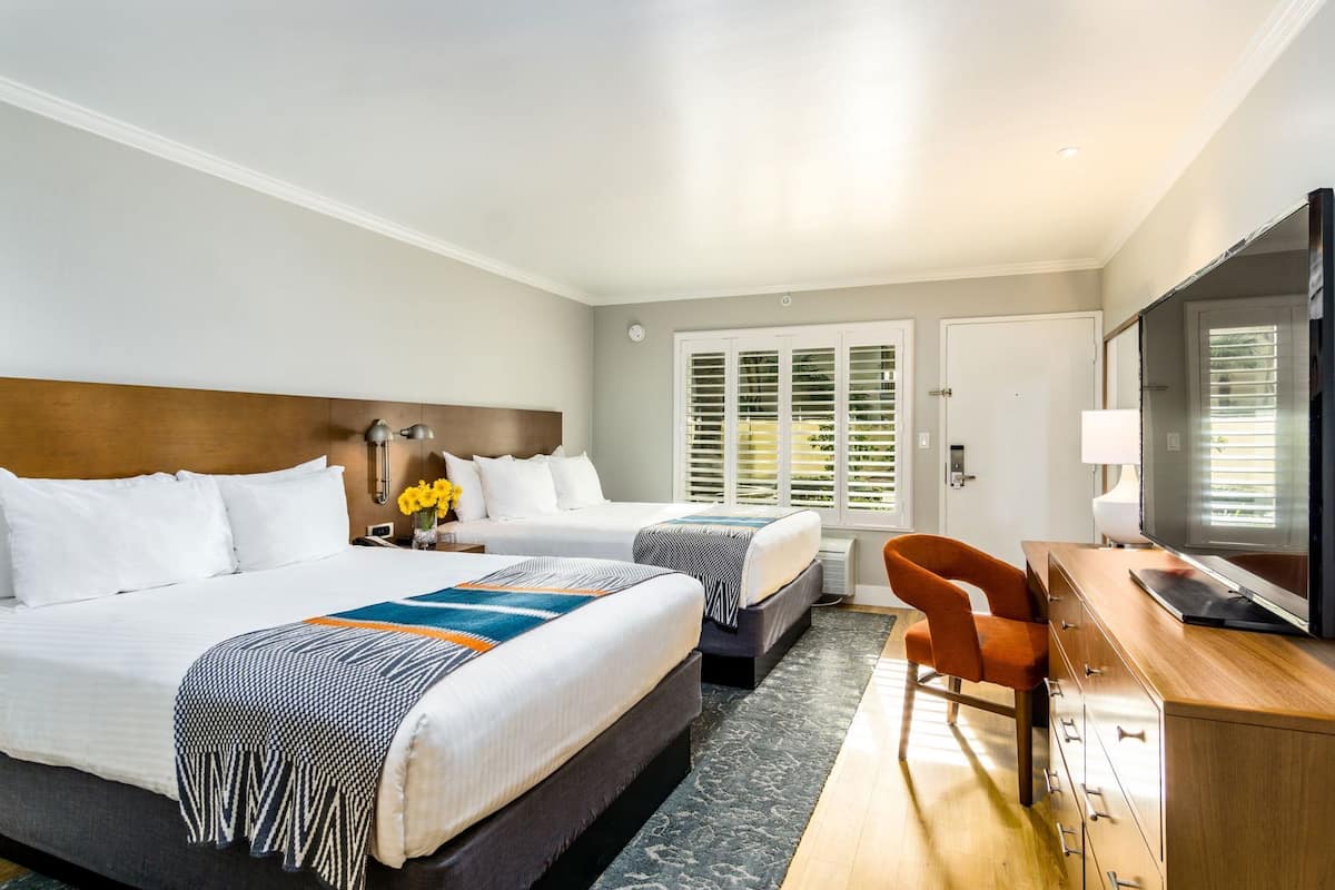 A hotel room with two beds, a desk, a television, and a window with blinds.