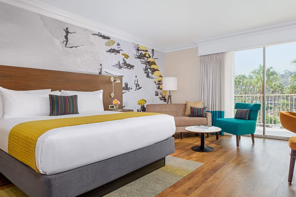 A hotel room with a large bed, a sofa, a teal armchair, and a balcony with a view of palm trees.