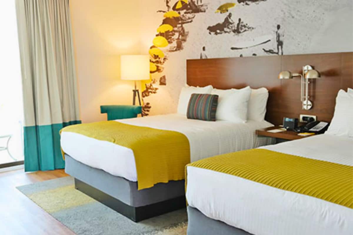 A hotel room with two beds, a desk, a chair, a lamp, and a view of the outdoors.