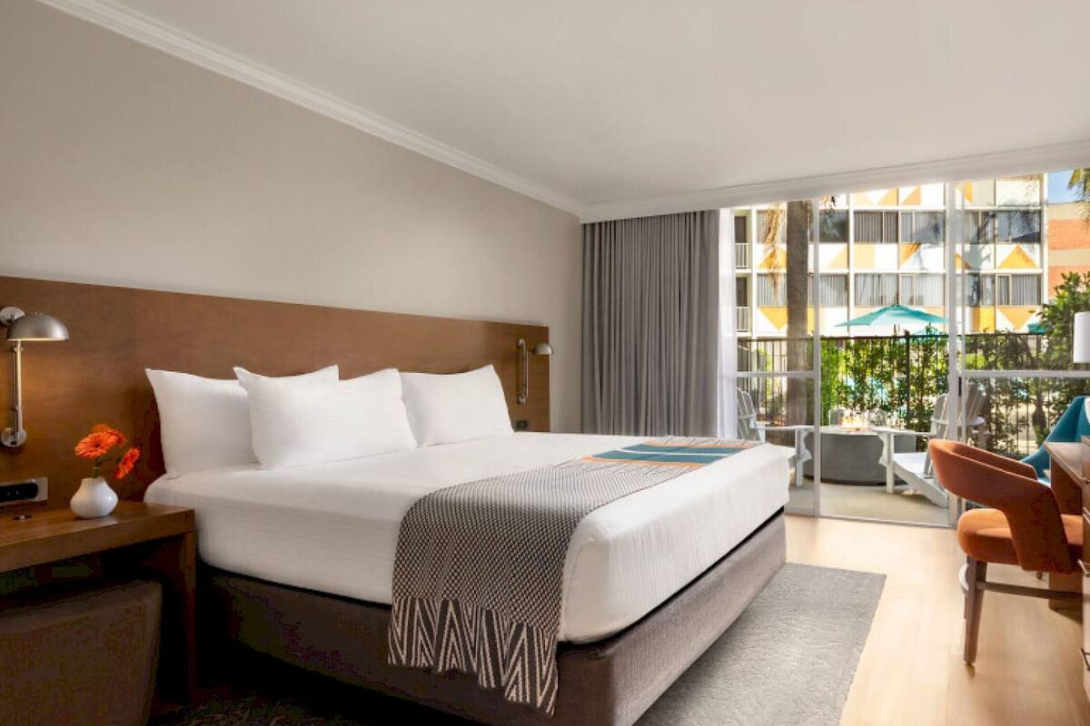 A hotel room with a large bed, a desk with a chair, a TV, and a view of the outside through a large window.
