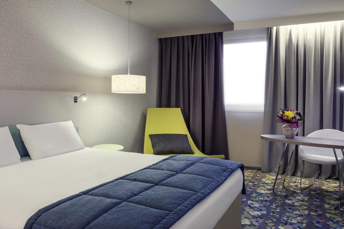 Une chambre dâhĂŽtel avec un lit, un bureau, une chaise, une lampe et une fenĂȘtre avec des rideaux.