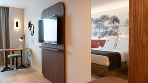 A modern hotel room with a flat-screen TV mounted on a wooden panel, a desk with a chair, a bed with white bedding, and a view of a snowy landscape through the window. - Mercure Paris Val de Fontenay (Fontenay-sous-Bois)