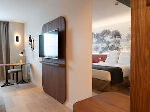 A modern hotel room with a flat-screen TV mounted on a wooden panel, a desk with a chair, a bed with white bedding, and a view of a snowy landscape through the window. - Mercure Paris Val de Fontenay (Fontenay-sous-Bois)