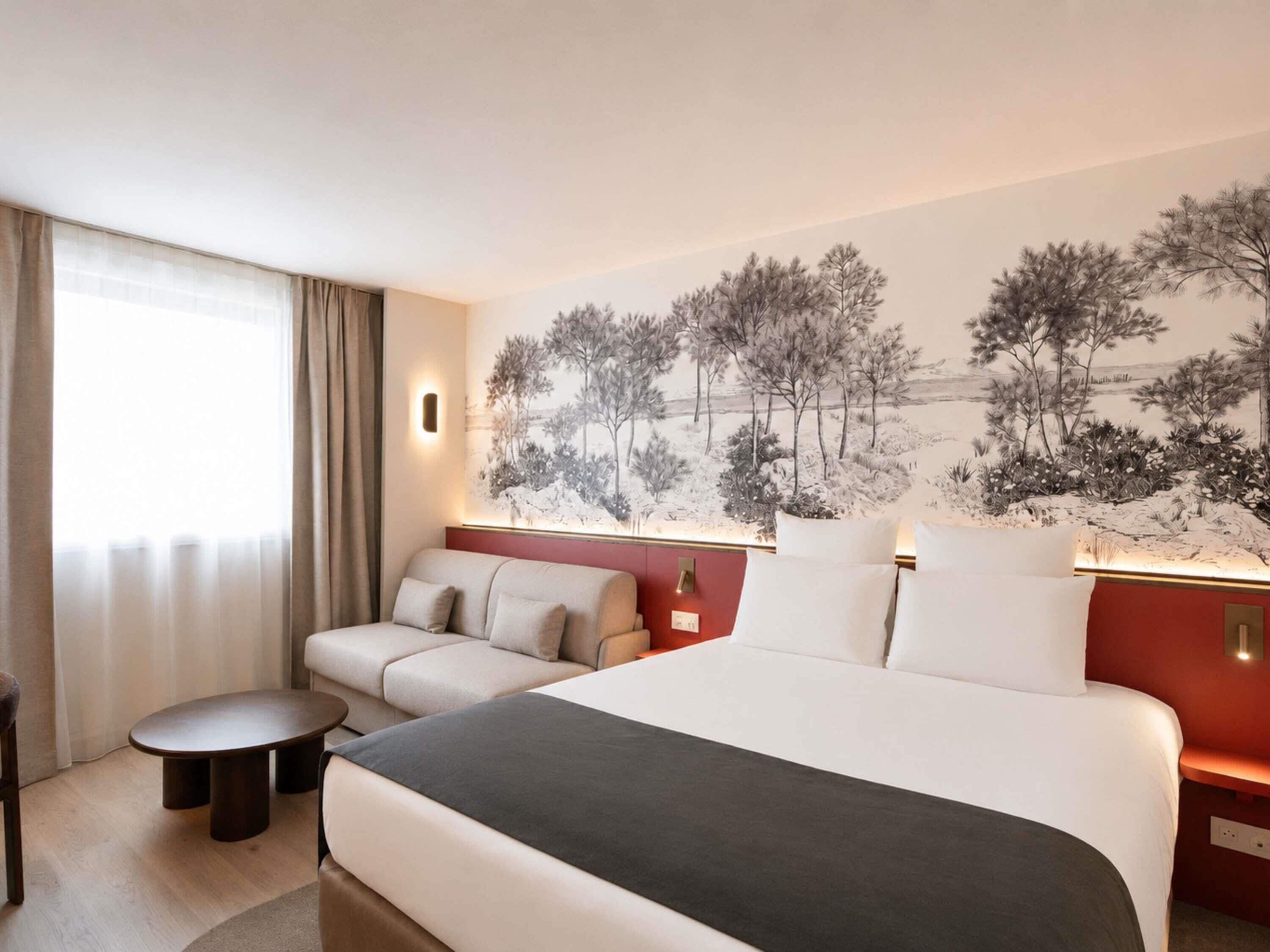 A hotel room with a bed, a sofa, a small table, and a wall-mounted lamp.