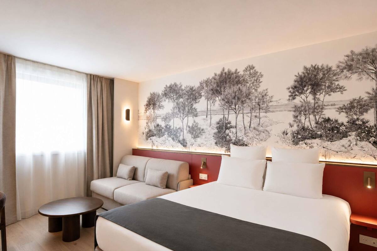 A hotel room with a bed, a sofa, a small table, and a wall-mounted lamp.