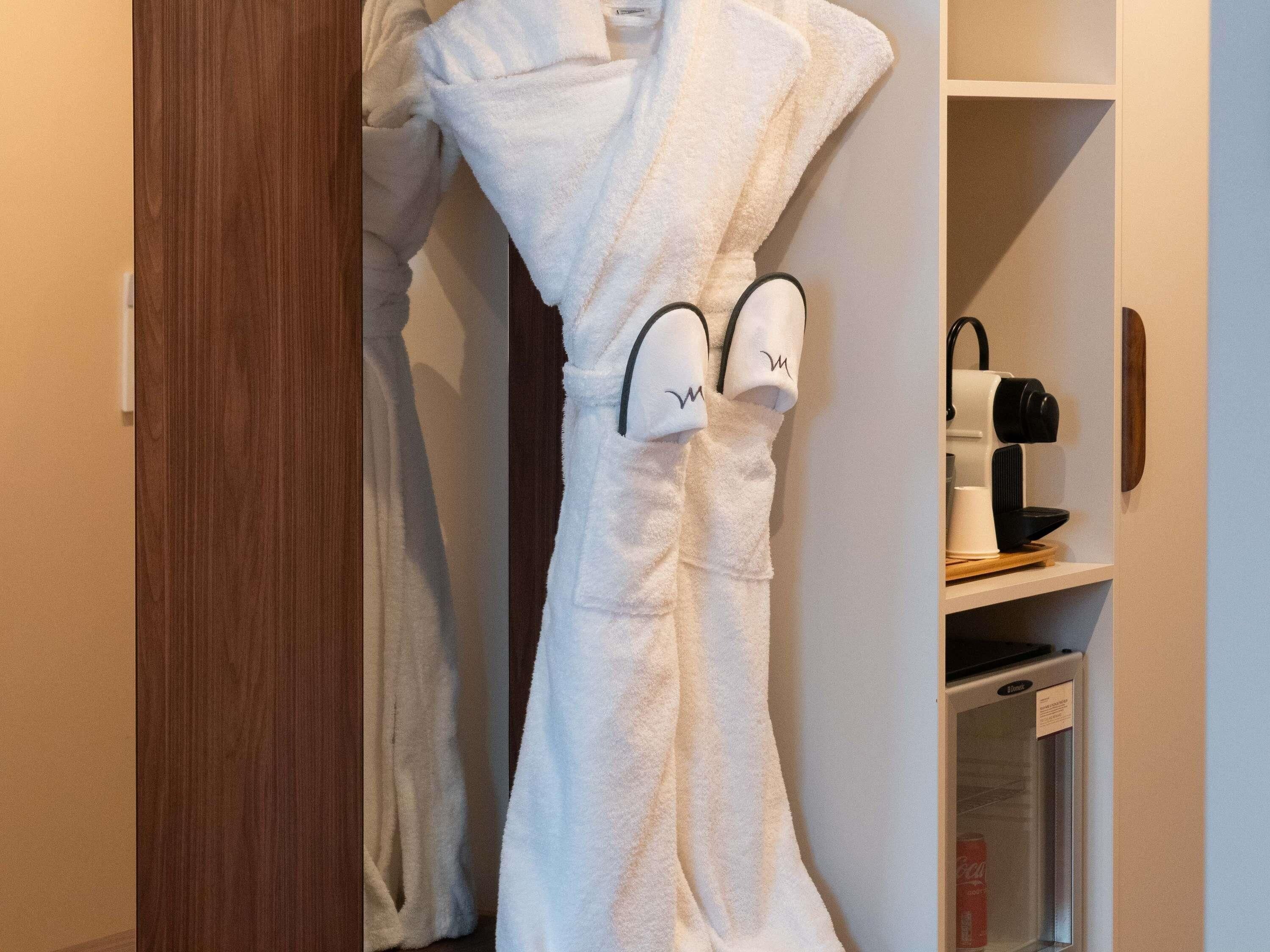 A white bathrobe hanging on a door, a coffee machine, and a refrigerator in a hotel room.