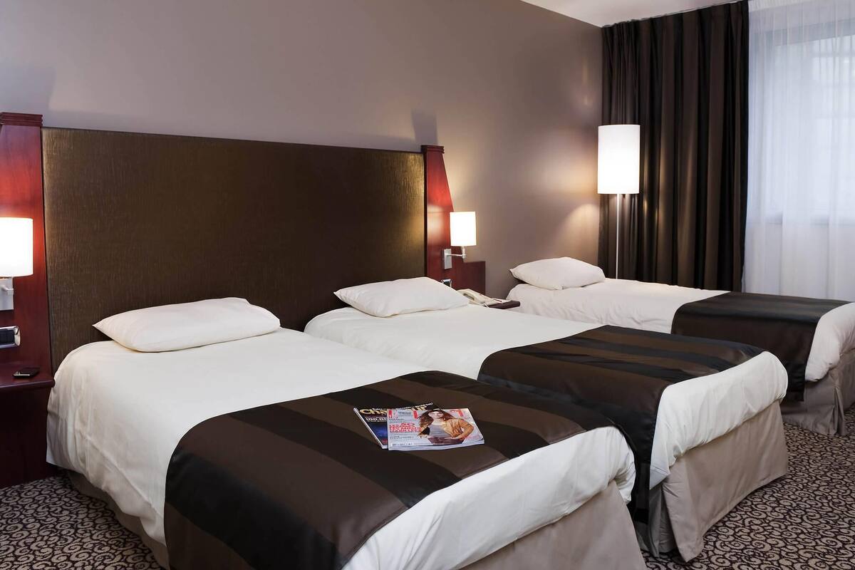 A hotel room with two beds, a nightstand with a lamp, a magazine on the bed, and a window with curtains.