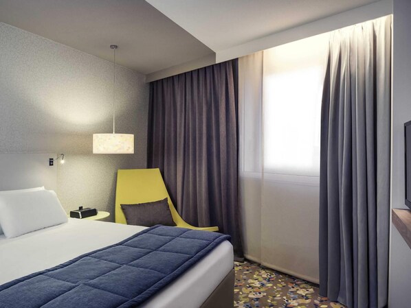A hotel room with a bed, a chair, a desk, a lamp, and a window with curtains.