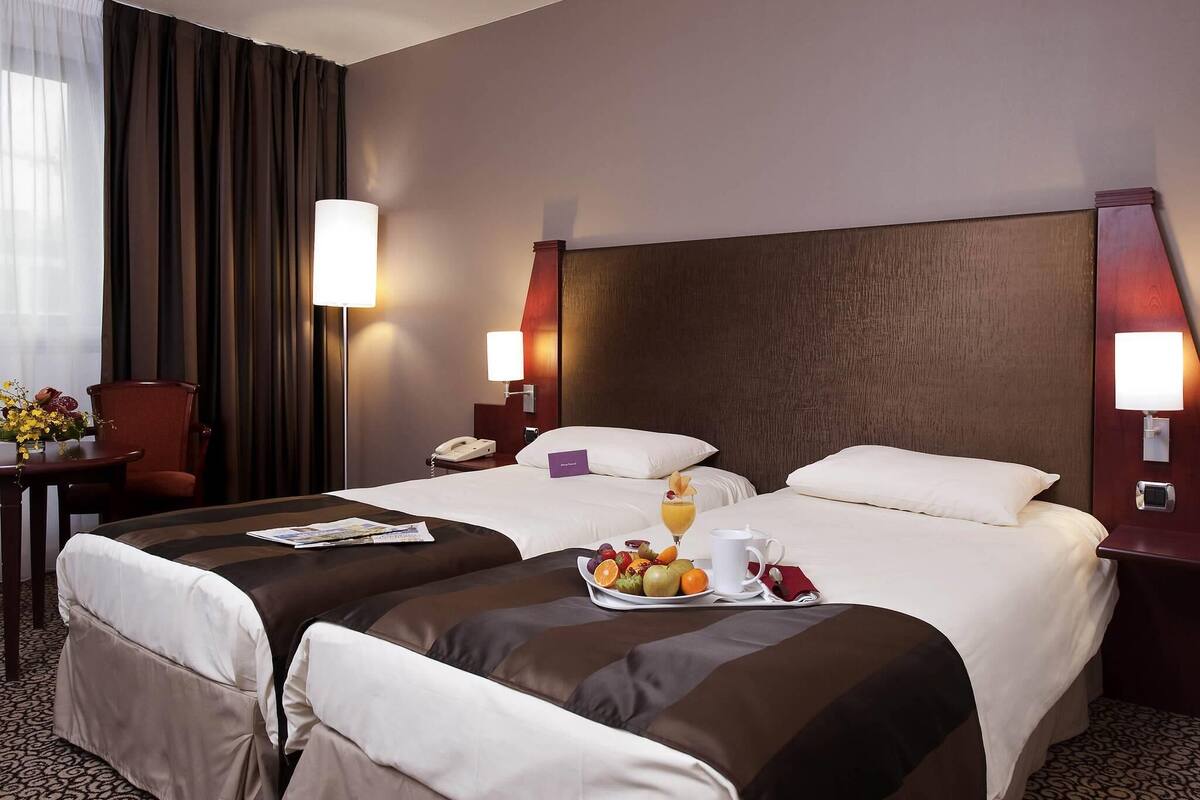 A hotel room with two beds, a desk, and a chair. There is a breakfast tray on the beds with fruits and a glass of orange juice.