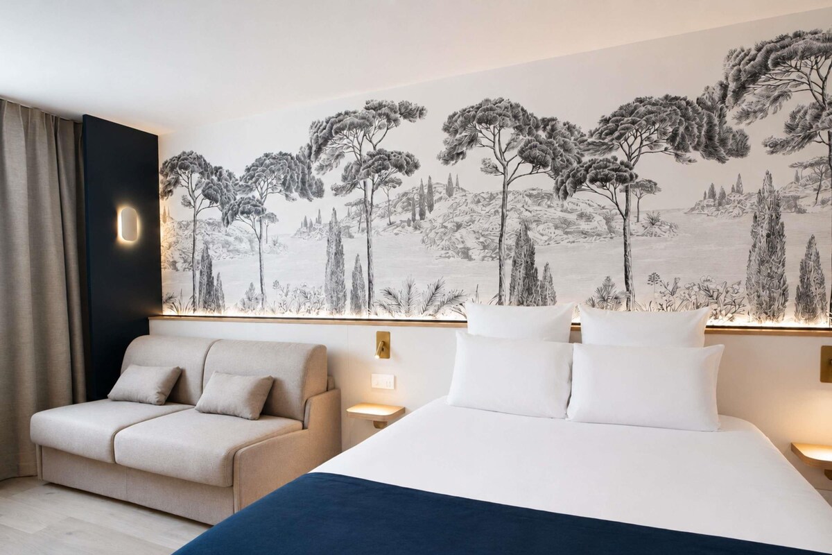 A hotel room with a bed, a sofa, a wall-mounted artwork, and bedside lamps.