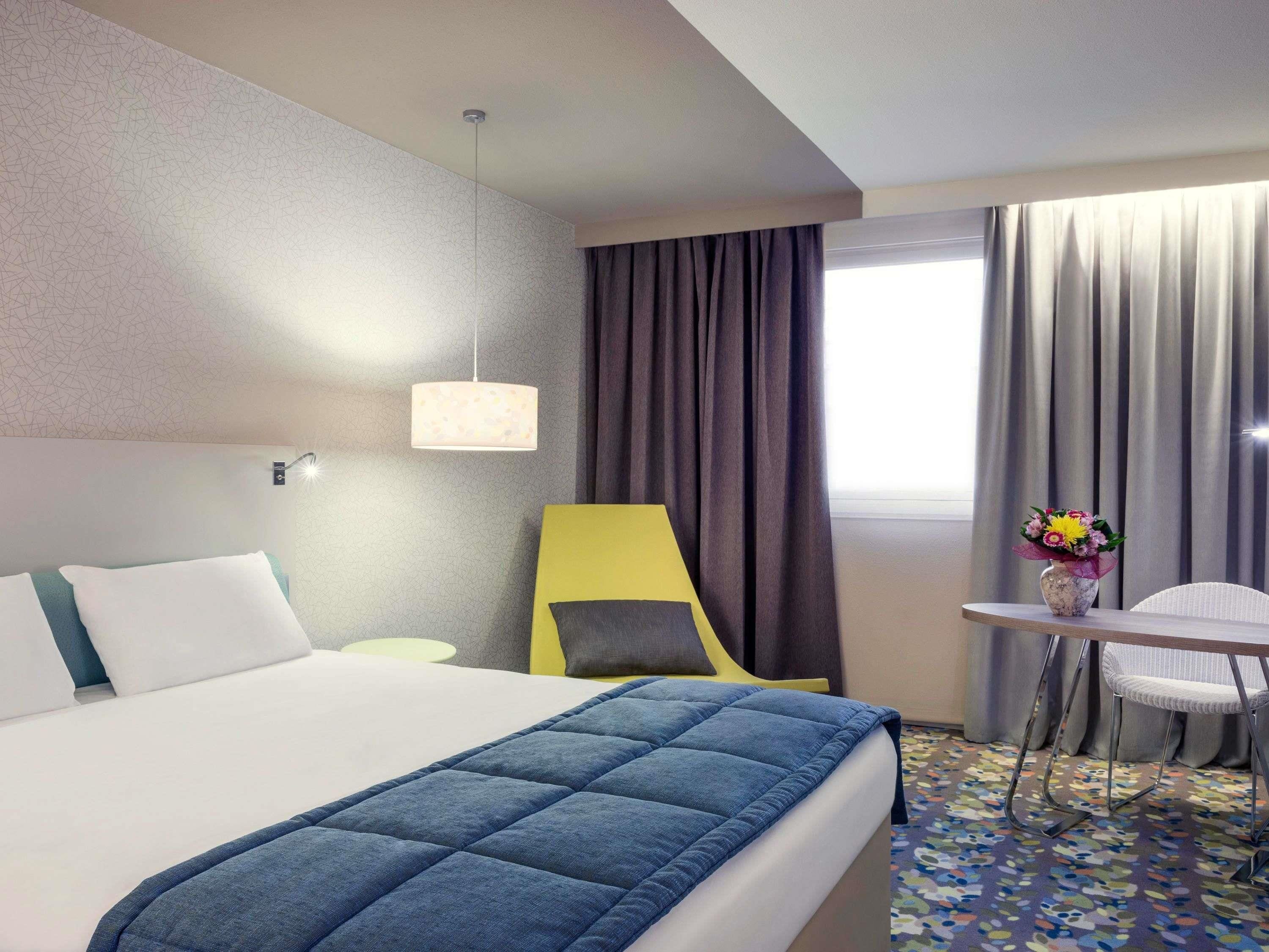 Ein Hotelzimmer mit Bett, Schreibtisch, Stuhl, Lampe und einem fenster mit Vorhängen.