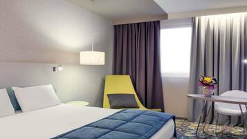 Ein Hotelzimmer mit Bett, Schreibtisch, Stuhl, Lampe und einem fenster mit Vorhängen.