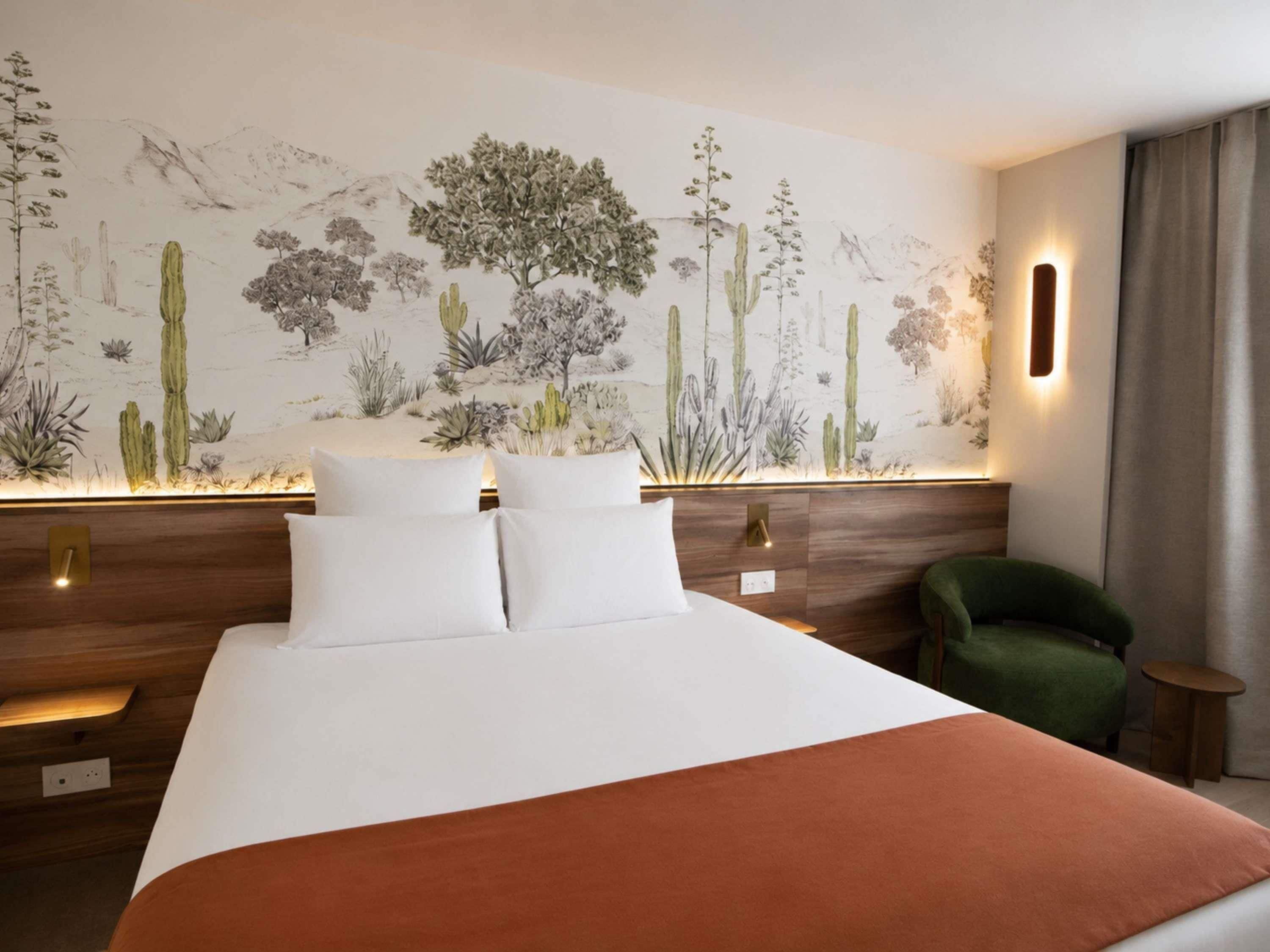 A hotel room with a large bed, a green armchair, a small table, and a wall mural of desert plants.