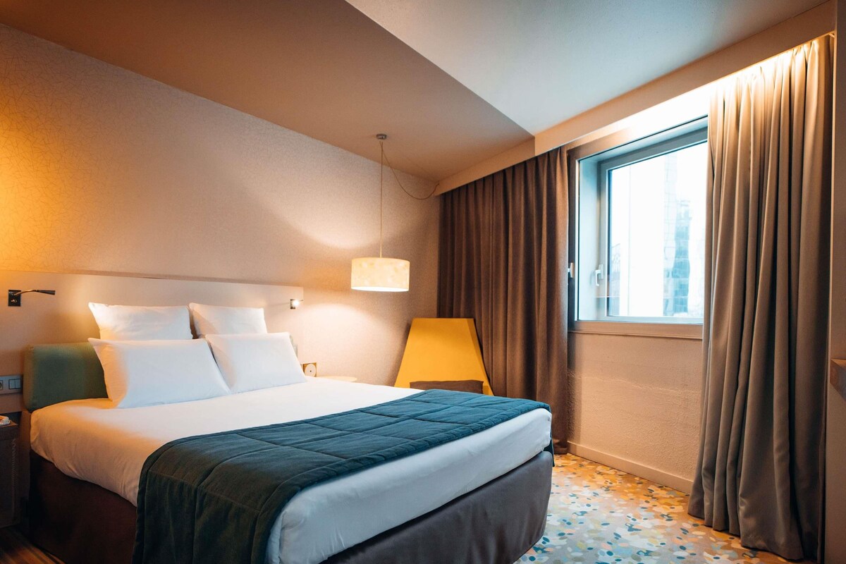 A hotel room with a bed, a nightstand, a lamp, a window with curtains, and a patterned floor.