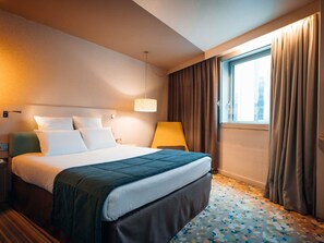 A hotel room with a bed, a nightstand, a lamp, a window with curtains, and a patterned floor. - Mercure Paris Val de Fontenay (Fontenay-sous-Bois)