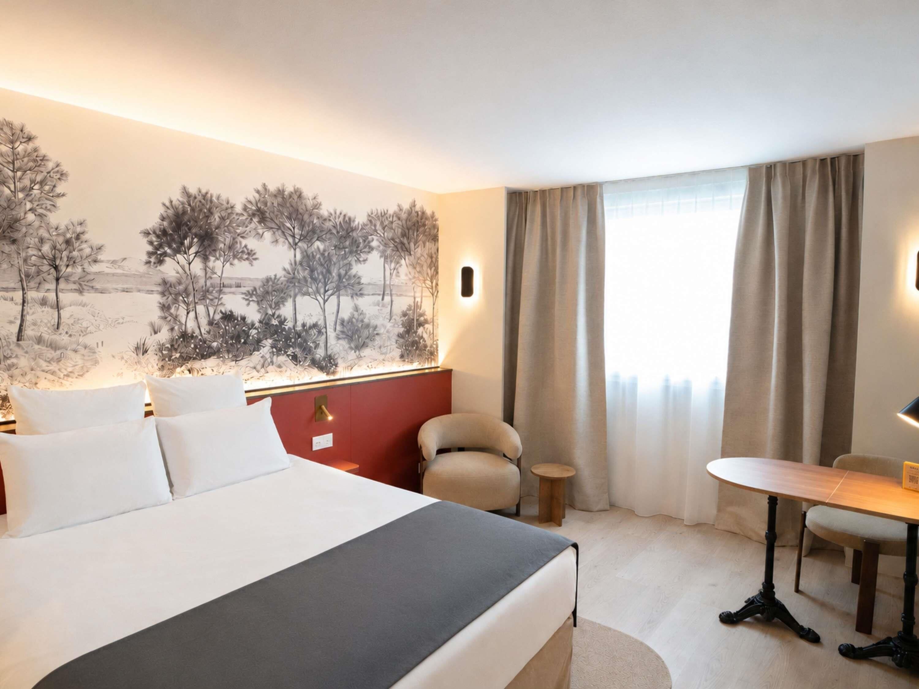 A hotel room with a bed, a desk, a chair, a window with curtains, and a wall mural.