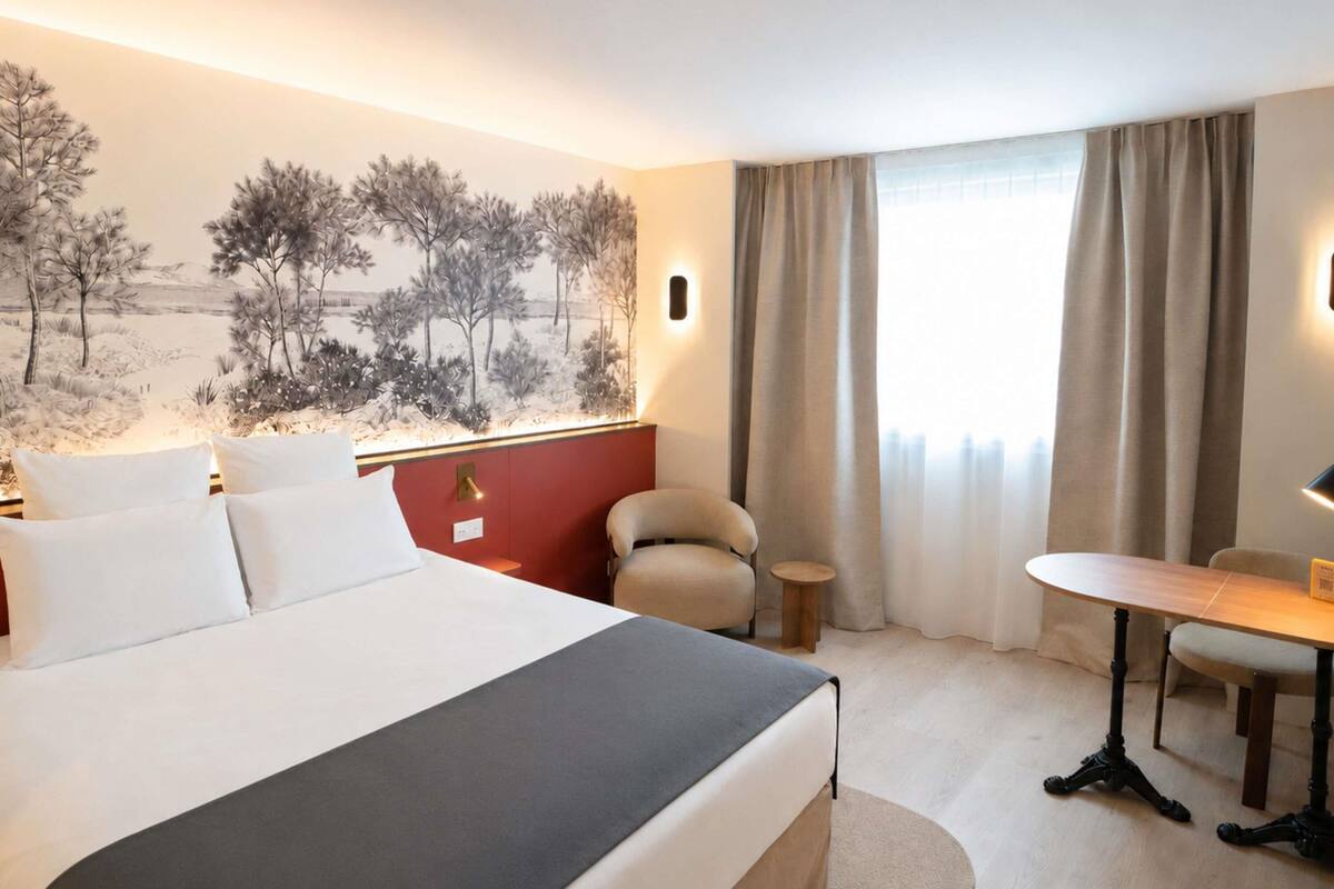 A hotel room with a bed, a desk, a chair, a window with curtains, and a wall mural.
