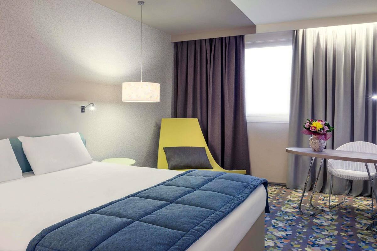 A hotel room with a bed, a desk, a chair, a lamp, and a window with curtains.