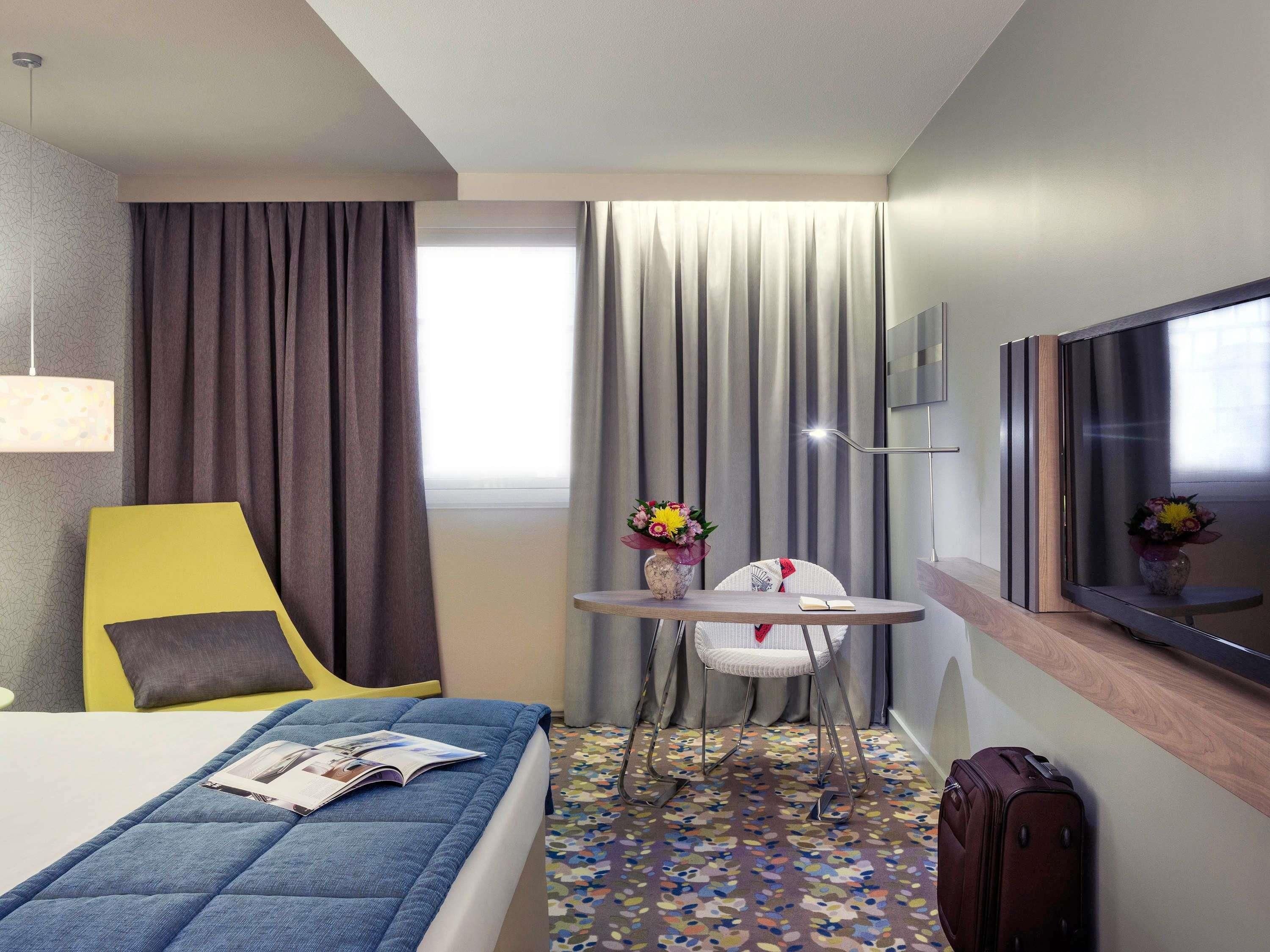 Une chambre d’hôtel avec un lit, une chaise, une petite table sur laquelle se trouve un vase, une télévision et une valise.